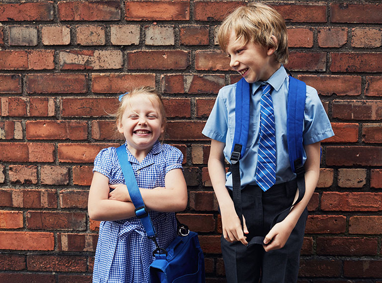 Two siblings in school uniforms smiling standing against a wall