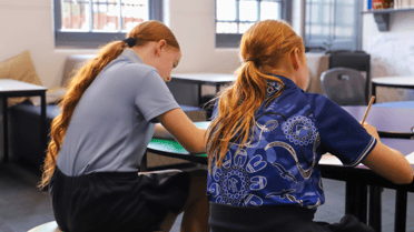 Two students sit at desks in a classroom, writing on paper during a lesson with tables, chairs and windows visible in the background.