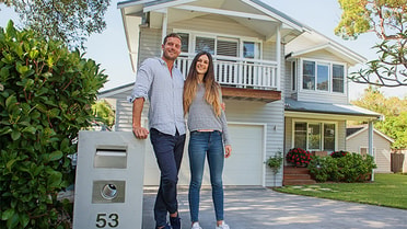 Bright man leans over post box and side hugs woman as they smile in front of a two storey house