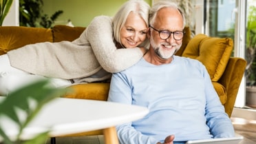 Older woman side lies on the couch as she leans on an older man sitting in front of couch as they smile at the laptop