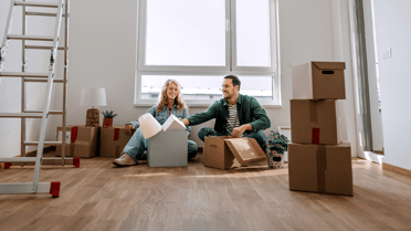 Two people sit on the floor of a new home surrounded by moving boxes, unpacking belongings in a bright room with large windows and a ladder nearby.