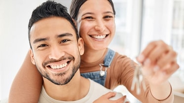 Two people pose indoors while holding up a set of house keys in the foreground, symbolising the purchase or move into a new home.