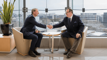 Two people seated across a small table shake hands in a modern office meeting area with floor-to-ceiling windows overlooking a city skyline.