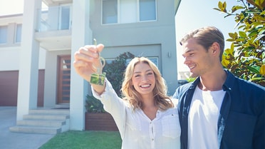 Young woman smiles holding up key while man interlinked smiles at her as they stand in front of a two storey modern house