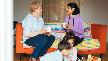 Two women sit in a couch with coffee cups in hand while children play games on the floor