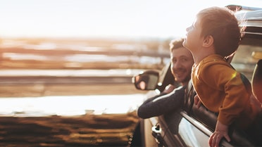 Father looks back at kid leaning out his window smiling in the back seat 