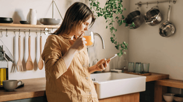 A woman drinking orange juice in her kitchen looking down at her phone